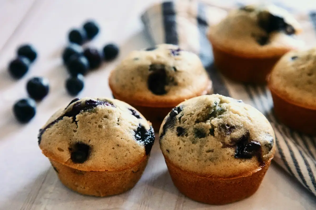 Freshly baked blueberry muffins with visible dark blueberries throughout, sitting on a wooden surface with a striped cloth and loose blueberries in the background.
