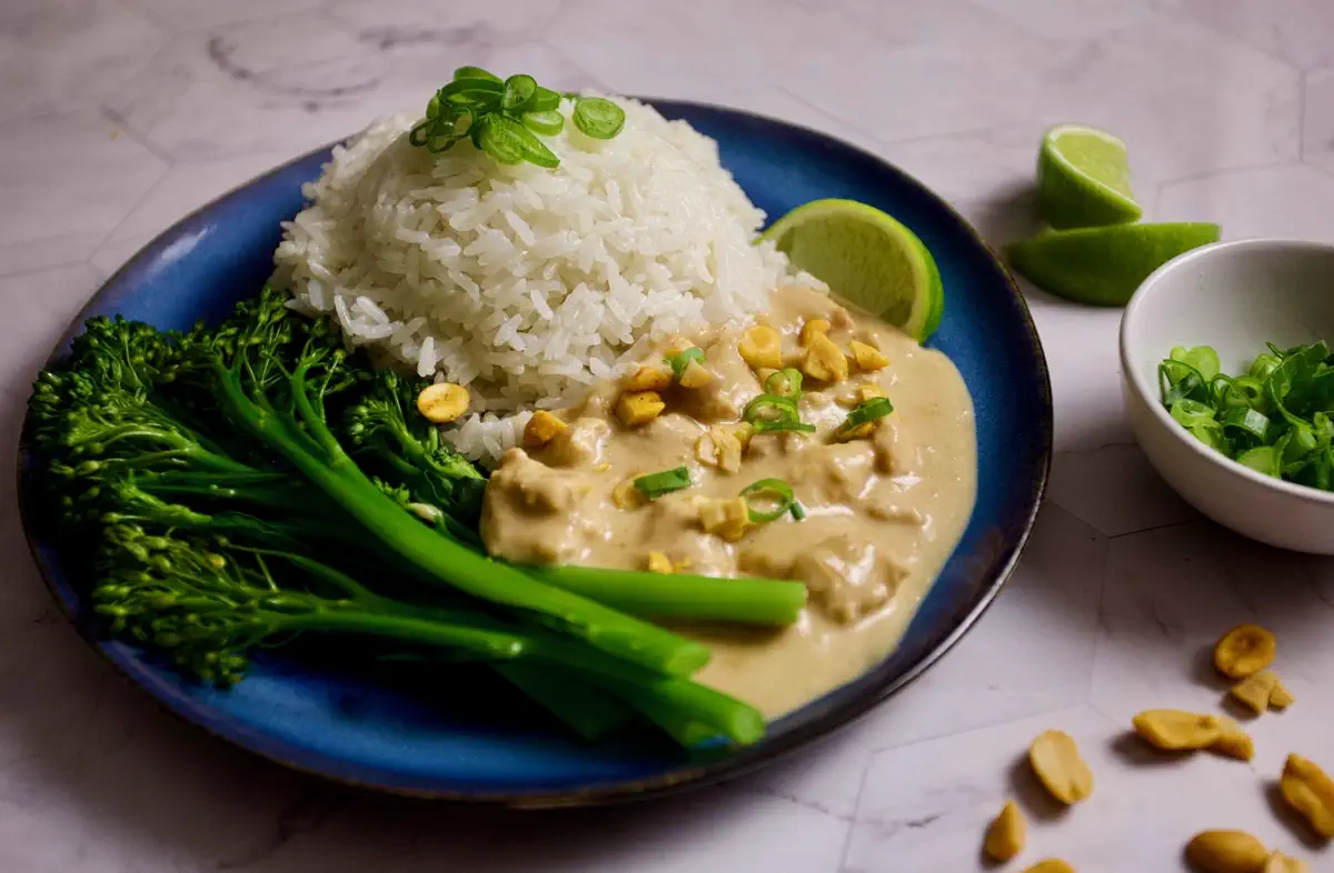 Chicken in a creamy peanut sauce served on a deep blue plate with fluffy jasmine rice and tender-stem broccoli, garnished with roasted peanuts, sliced spring onions, and a wedge of lime.