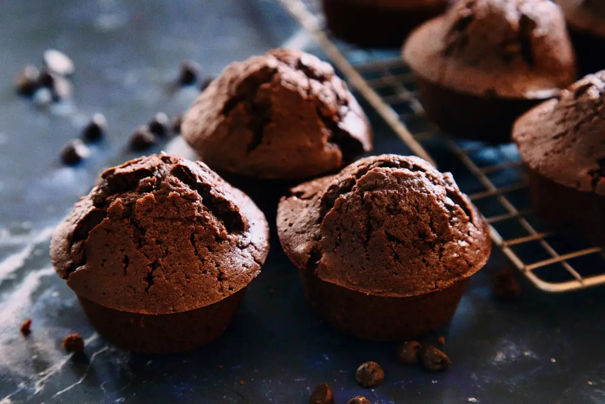 Rich chocolate muffins with visible dark chocolate chips, unmoulded from a tin and cooling on a metal rack, with extra chocolate chips scattered on the work surface.