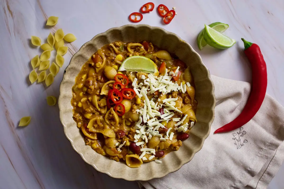 Bowl of cheesy taco pasta with beef, pasta shells, corn, beans, fresh chili slices, and lime on a light background.