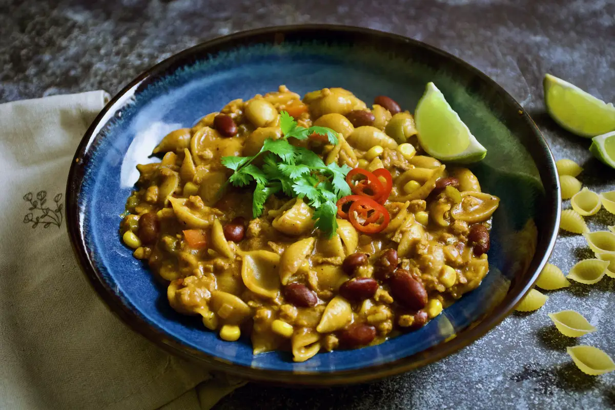 Bowl of cheesy vegan taco pasta with plant-based mince, pasta shells, corn, beans, fresh chili slices, and lime on a light background.